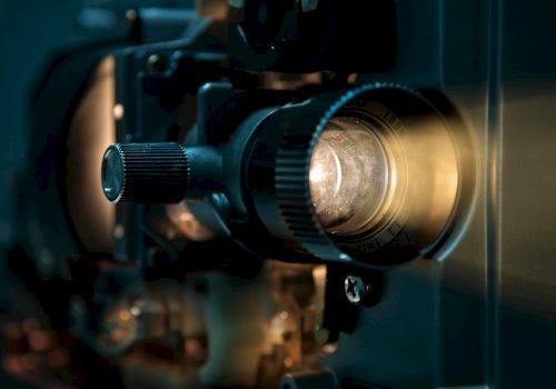 Close-up of a teal camera/projector lens with focus markings and glowing glass, mounted on a mechanical device.