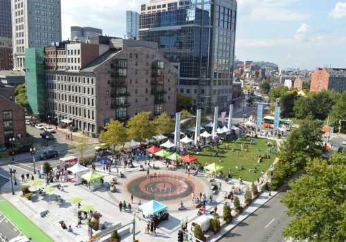 A sunny city plaza packed with people, green tents, food stalls, and a central circular fountain amid tall buildings.