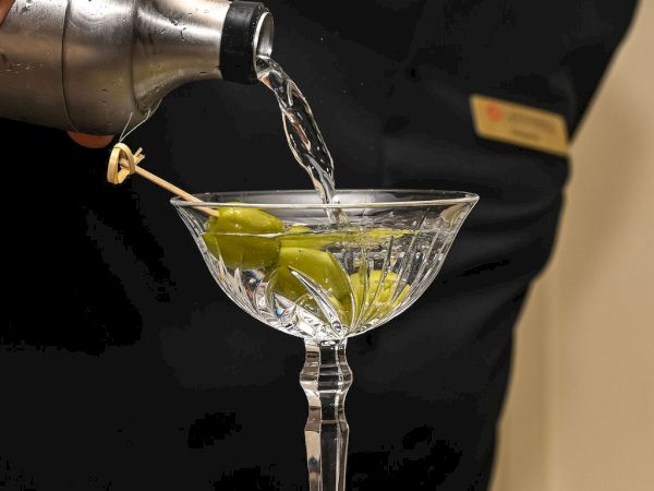 A bartender pours a clear drink into a cocktail glass with green garnishes on the bar, for a fresh serve.