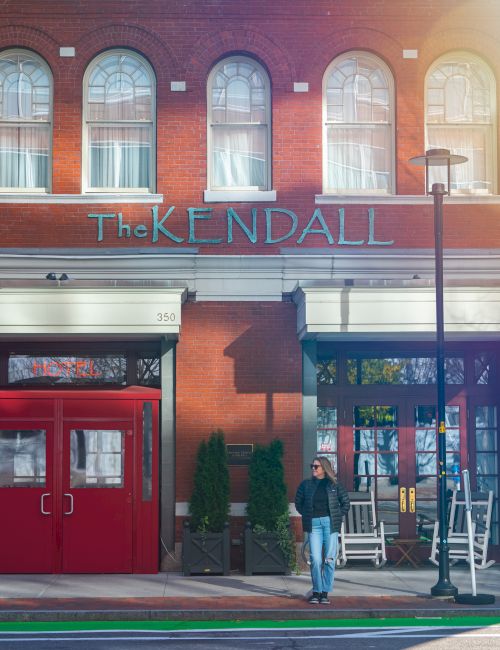 A brick hotel building with large windows and the sign &ldquo;The Kendall,&rdquo; with a person standing outside.