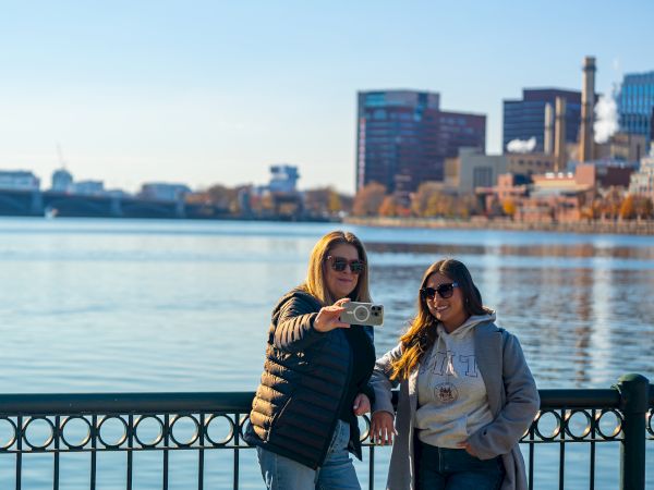 Two friends smile for a selfie by a river, with a city skyline and calm water in the background, soaking up the sunshine.