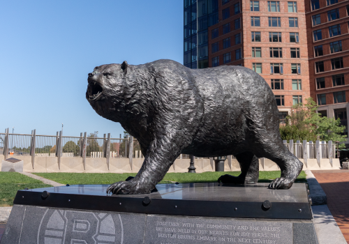 A bronze bear statue stands on a plaza in front of tall office buildings under a clear blue sky.
