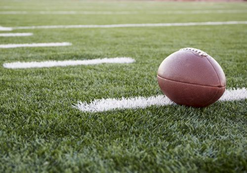 A brown American football rests on green artificial turf near the yard line, ready for the next play.