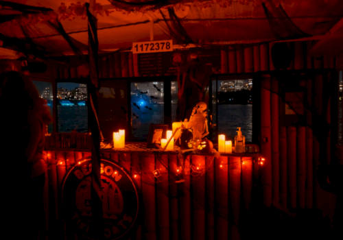 Interior of a vessel at night, lit with red candles and incense around a small statue, with city lights through the windows.
