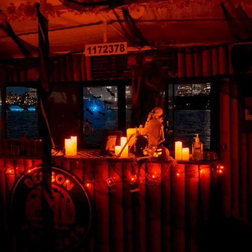 Interior of a vessel at night, lit with red candles and incense around a small statue, with city lights through the windows.