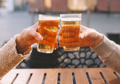 Two people clink amber beer glasses, toasting with frothy drinks outdoors in bright sunshine.
