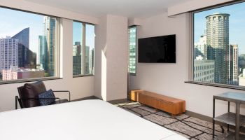 This image shows a modern hotel room with large windows offering a city skyline view, a bed, a TV, a chair, and a table.
