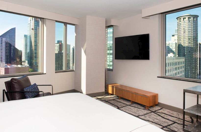 This image shows a modern hotel room with large windows offering a city skyline view, a bed, a TV, a chair, and a table.