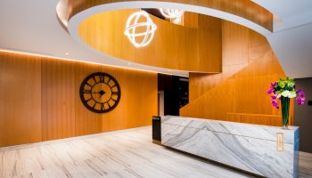 A modern lobby with a curved wooden staircase, a large wall clock, a marble reception desk, and a flower arrangement.