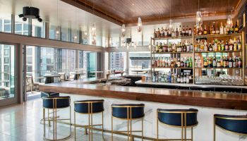 The image shows a modern bar with chic barstools, an extensive liquor display, and a view of a cityscape through large windows, ending the sentence.