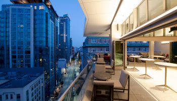 The image shows a modern cityscape at dusk with high-rise buildings, and a spacious balcony with outdoor seating and tables.