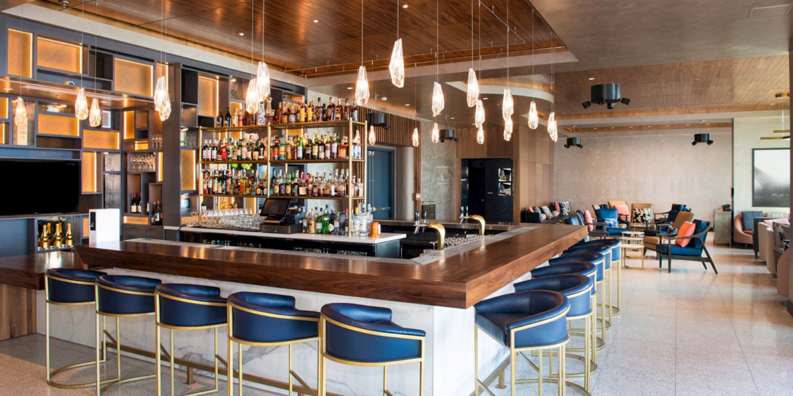 The image shows a modern bar with a central wooden counter, blue bar stools, various liquors on display, and pendant lights overhead.