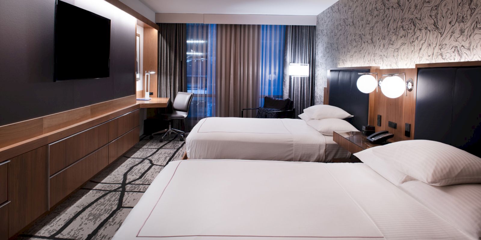 A hotel room with two double beds, a TV, a desk with a chair, and modern decor and lighting.