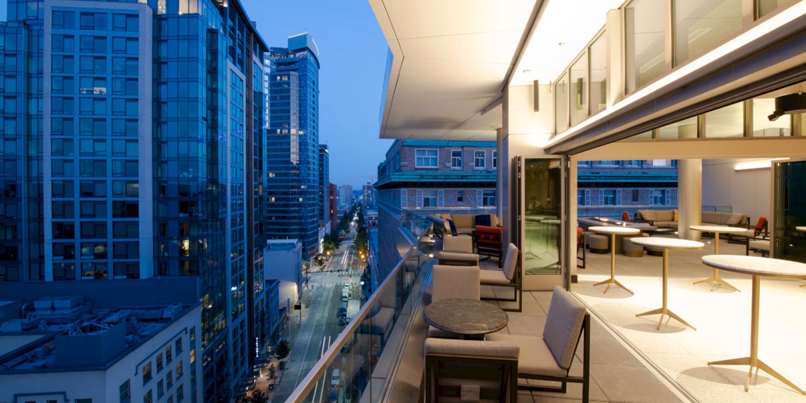 The image shows a modern balcony with seating, overlooking an urban cityscape with tall buildings at dusk.