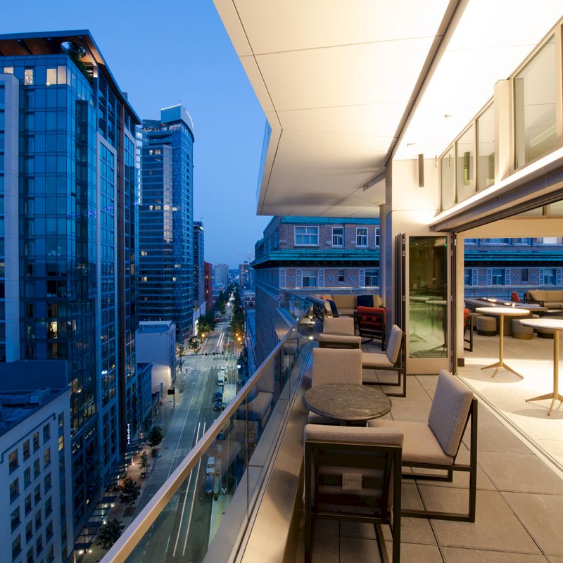 A modern balcony view with a cityscape at twilight, featuring outdoor seating, glass railings, tall buildings, and a street below.