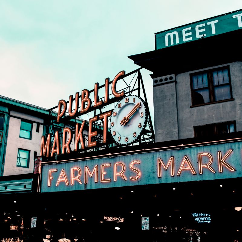 A large neon sign reads "Public Market" and "Farmers Market" in front of commercial buildings, with a clock displayed prominently in the center.