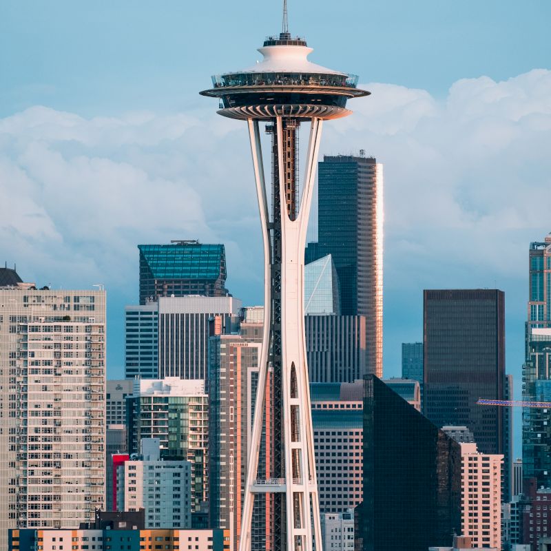This image features the iconic Space Needle in Seattle, Washington, surrounded by modern skyscrapers and urban buildings under a partly cloudy sky.