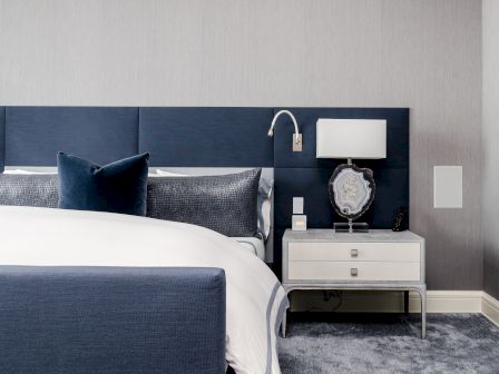 A modern bedroom featuring a blue headboard, white bedside table, table lamp, and dark blue bedding with pillows, all on a gray carpet.