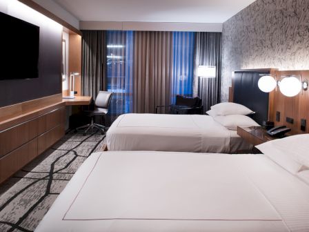 A modern hotel room with two double beds, a TV mounted on the wall, a desk with a chair, ambient lighting, and large windows with curtains.