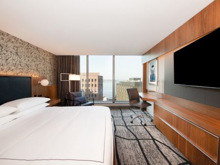A modern hotel room with a large bed, wall-mounted TV, desk, and a chair. There's a window with a view of buildings and water outside.