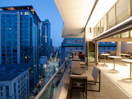 This image shows a modern balcony overlooking a city street with tall buildings at twilight, featuring tables, chairs, and open windows.