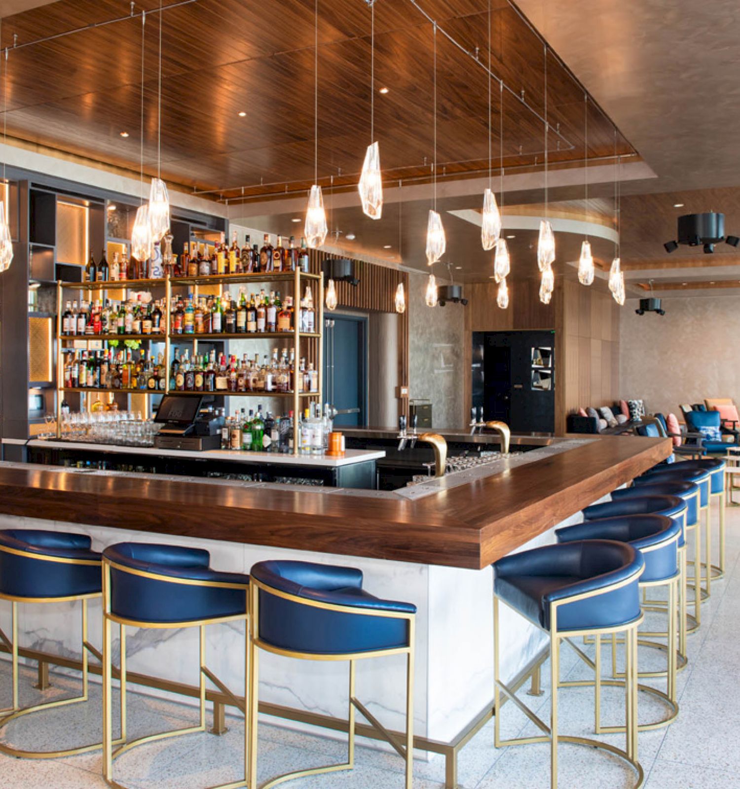 A modern bar with a wooden countertop, blue stools, hanging lights, and a well-stocked liquor shelf in a spacious, stylish interior.