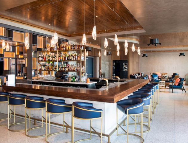 The image shows a modern bar with blue barstools, a wooden counter, hanging lights, and a well-stocked liquor shelf.