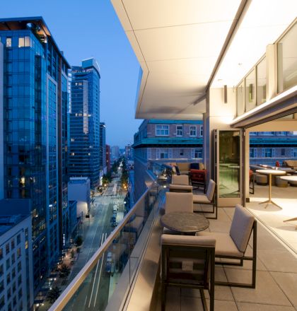 The image shows a modern cityscape with tall buildings at evening time, viewed from a balcony with seating and tables on the right side.