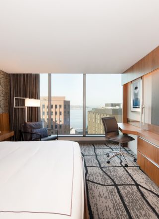 A modern hotel room with a large bed, TV, desk, and chair, featuring a city view through floor-to-ceiling windows, ending the sentence.
