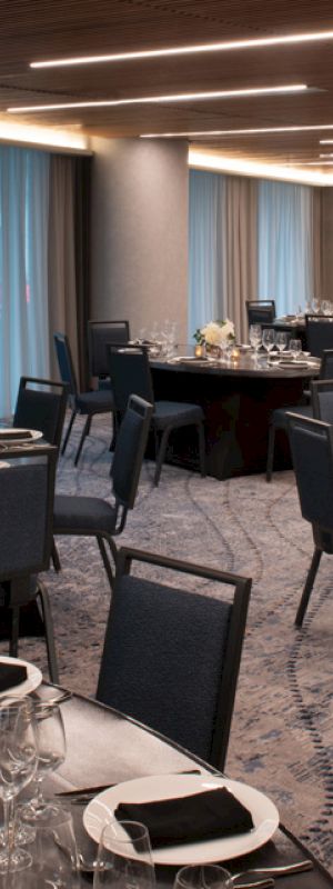 The image shows a modern, elegantly set dining room with round tables, black chairs, white floral centerpieces, and dim lighting for an event.