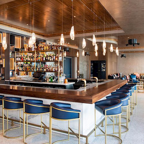 A modern bar with a wooden countertop, blue cushioned stools, a variety of drinks on shelves, stylish lighting, and a seating area in the background.