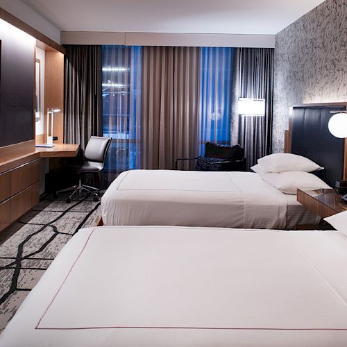 A modern hotel room with two double beds, a flat-screen TV, a work desk with a chair, and a large window with curtains.