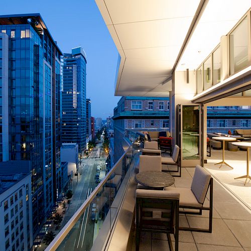 A modern urban balcony at twilight, with outdoor seating and tables, overlooks a city street lined with tall buildings.