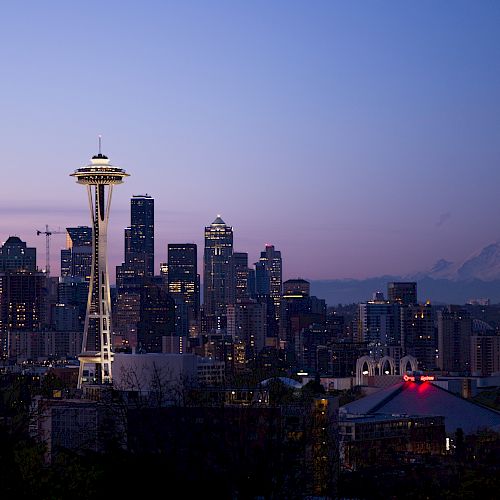 A twilight cityscape with a prominent needle-like tower and a silhouetted mountain in the background, with buildings lit up against the evening sky.