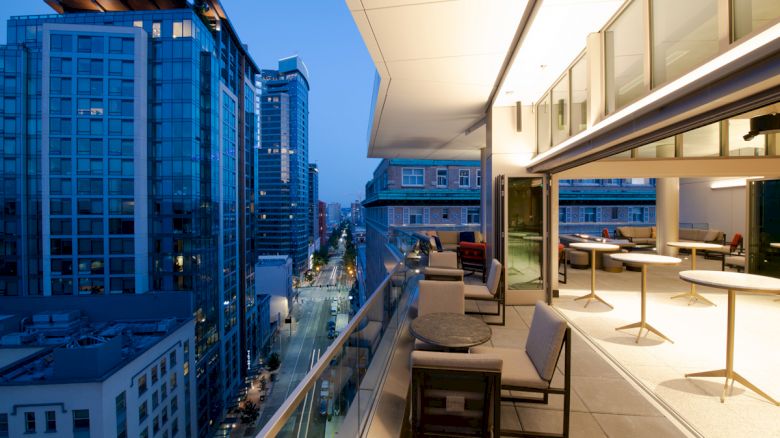 A modern rooftop patio with tables and chairs overlooks a city street lined with tall buildings at dusk, with a brightly lit interior space nearby.