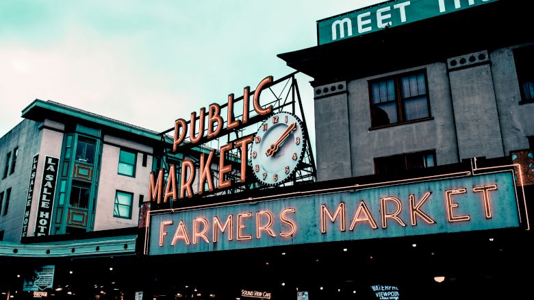 Colorful neon signs for "Public Market" and "Farmers Market" on buildings, with a street clock beneath an evening sky.