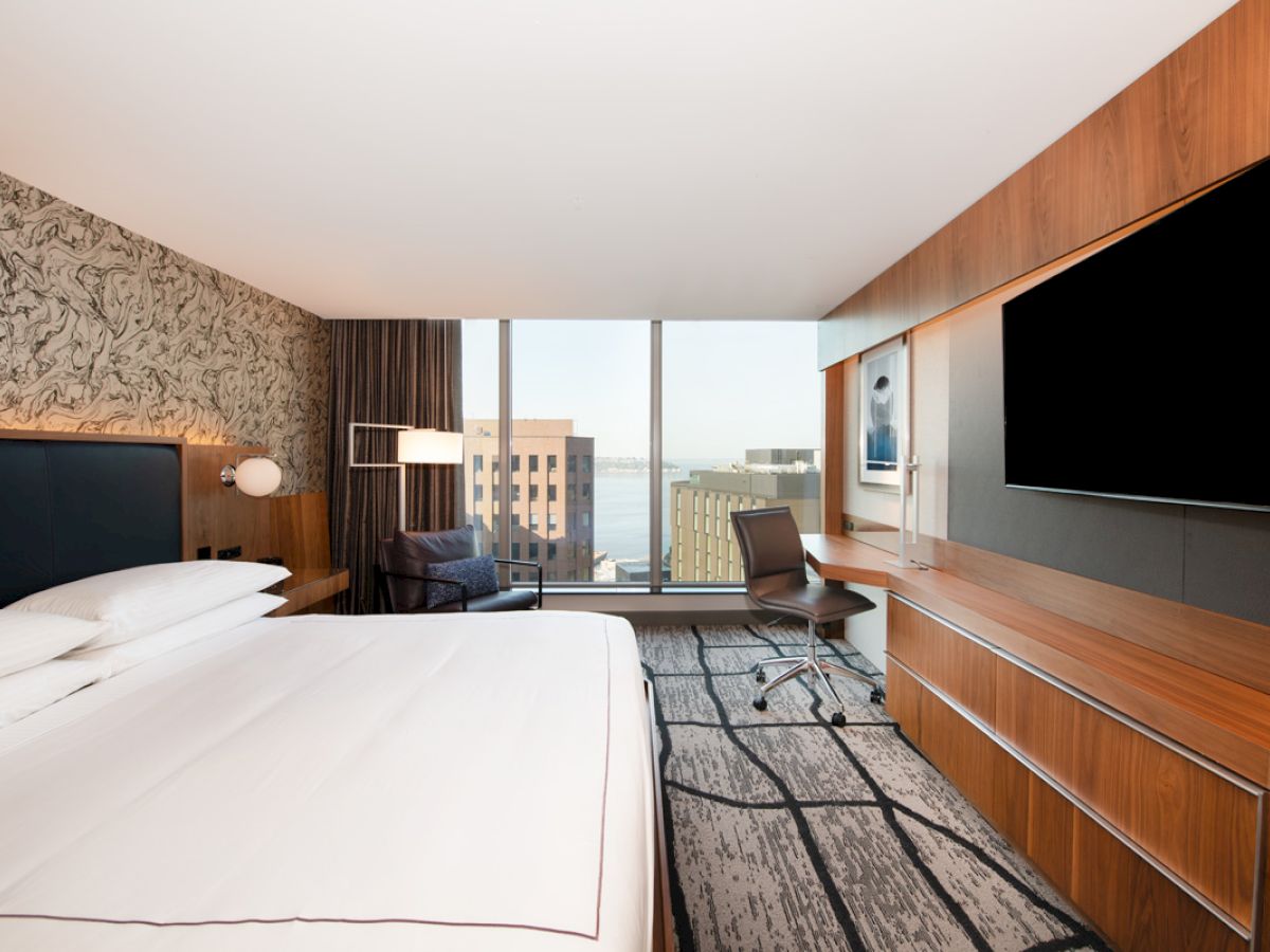 A modern hotel room with a large bed, flat-screen TV, desk, chair, and a city view through a window.