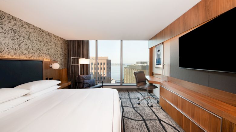 A modern hotel room with a large bed, flat-screen TV, desk, chair, and a city view through a window.