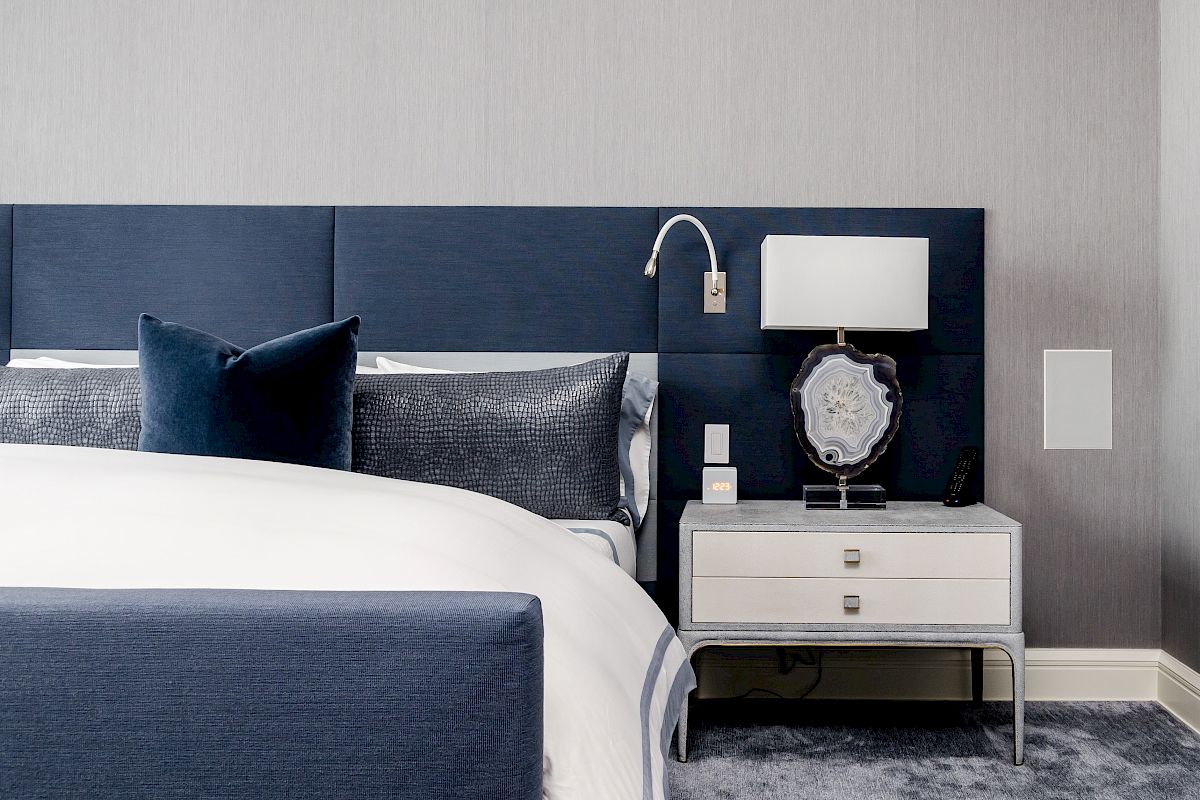 A modern bedroom features a blue and white color scheme with a padded headboard, nightstand, lamp, and decorative pillows on a well-made bed.