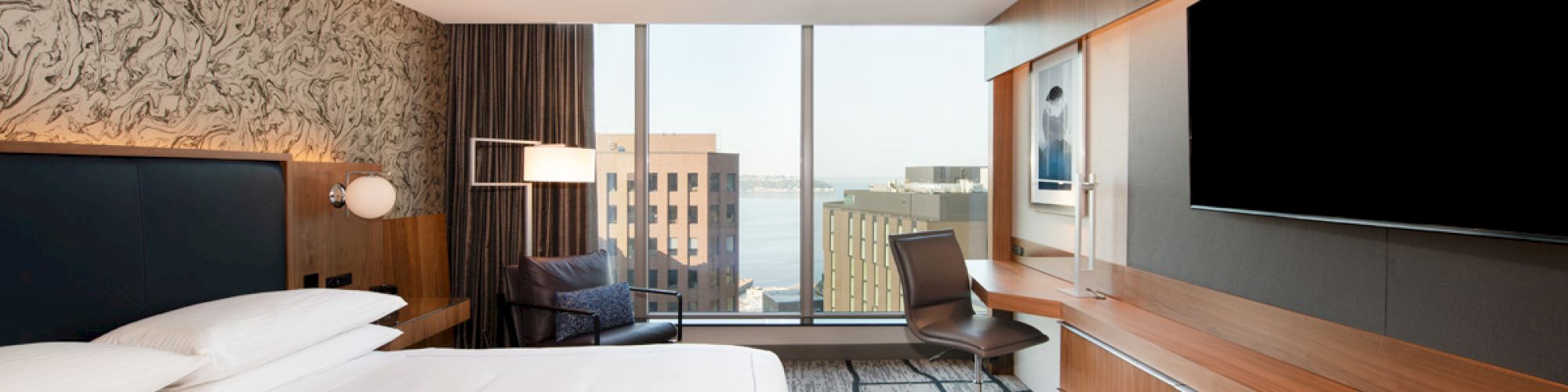 A modern hotel room features a large bed, a flat-screen TV, a work desk, and a seating area next to a window with a view of city buildings.